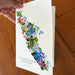 A hand holds a white greeting card featuring a colorful floral illustration of Vancouver Island, Canada, against a warm wooden tabletop background.
