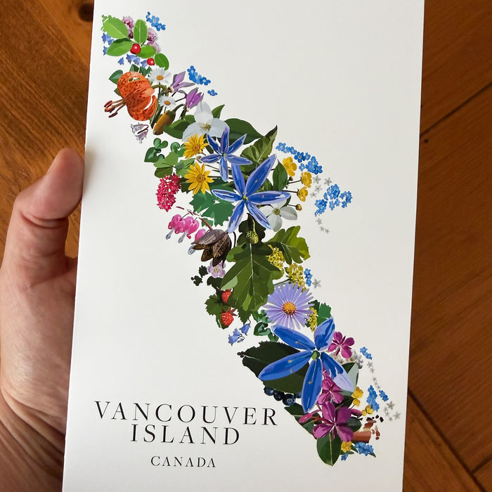A hand holds a white botanical art greeting card depicting the map of Vancouver Island filled with colorful wildflowers and green foliage, with the text 'VANCOUVER ISLAND CANADA' at the bottom, set against a wooden floor background.
