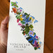 A hand holds a white botanical art greeting card depicting the map of Vancouver Island filled with colorful wildflowers and green foliage, with the text 'VANCOUVER ISLAND CANADA' at the bottom, set against a wooden floor background.