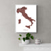 Framed map of Italy on a wall above a white shelf with a plant and mug.