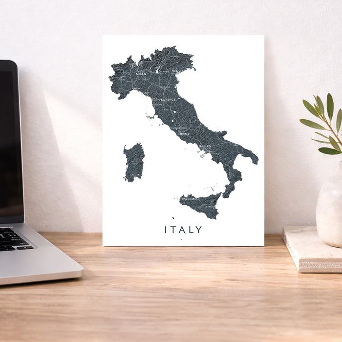 Map of Italy on a white background next to a laptop on a wooden desk.