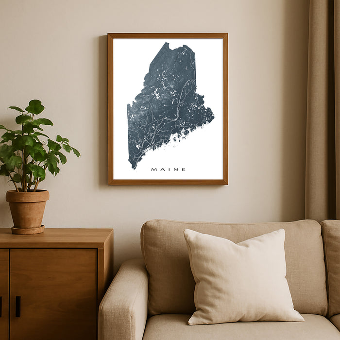 Framed map of Maine on a wall above a beige sofa with a plant beside it.
