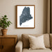 Framed map of Maine on a wall above a beige sofa with a plant beside it.