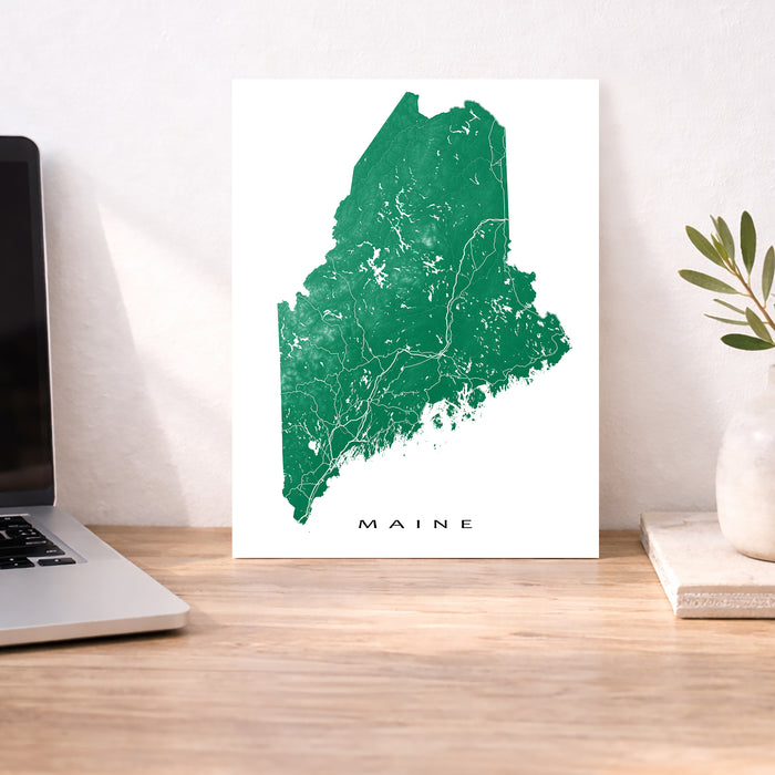 Map of Maine on a white background next to a laptop on a wooden desk.