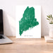 Map of Maine on a white background next to a laptop on a wooden desk.