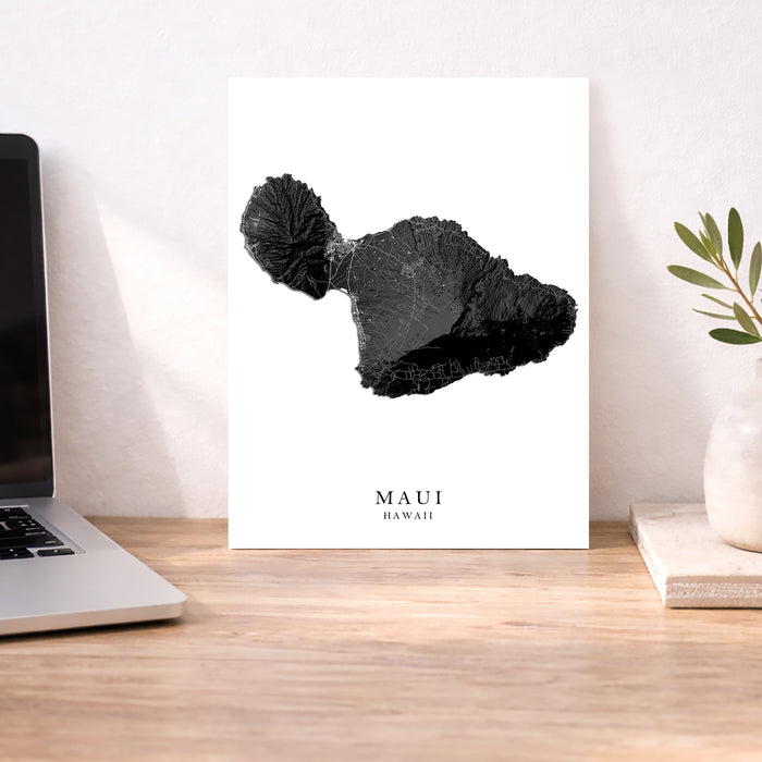 Art print of black map with 'MAUI HAWAII' text on a desk next to a laptop.