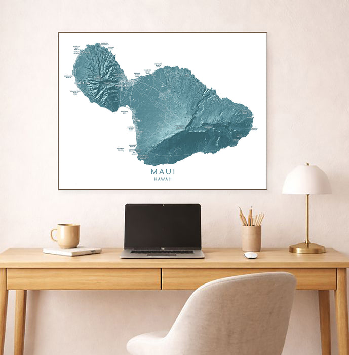 Framed map of Maui, Hawaii on a wall above a desk with a laptop and lamp.