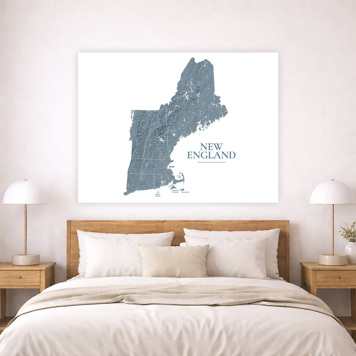 A large square wall art print showing a blue-gray map of New England on a white background, displayed above a neatly made bed in a bright bedroom with two bedside lamps.