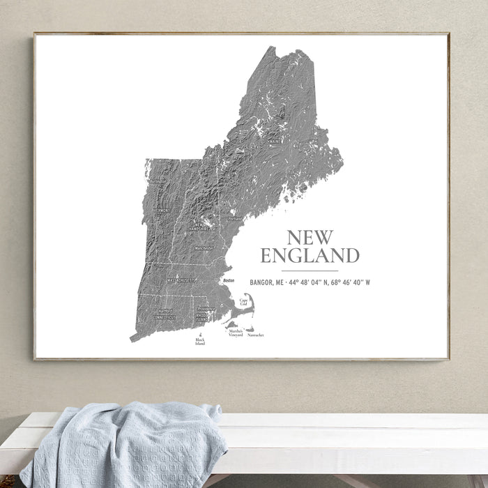 A framed wall art print showing a gray topographic map of New England on a white background, hanging on a beige wall above a white bench with a light blue cloth draped over it.