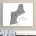 A framed wall art print showing a gray topographic map of New England on a white background, hanging on a beige wall above a white bench with a light blue cloth draped over it.