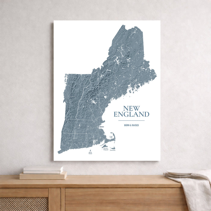 A vertical wall art print featuring a blue-gray textured map of New England with the words 'NEW ENGLAND' and 'BORN & BAGED' on a white background, displayed above a wooden console table in a styled interior setting.
