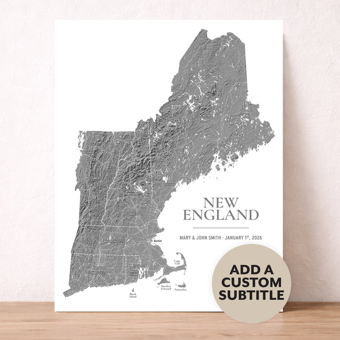 A custom topographic map print of New England, featuring a gray shaded relief map of the region on a white background, with the text 'NEW ENGLAND' and a customizable subtitle. A beige circular badge in the bottom right corner reads 'ADD A CUSTOM SUBTITLE'.
