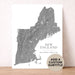 A custom topographic map print of New England, featuring a gray shaded relief map of the region on a white background, with the text 'NEW ENGLAND' and a customizable subtitle. A beige circular badge in the bottom right corner reads 'ADD A CUSTOM SUBTITLE'.