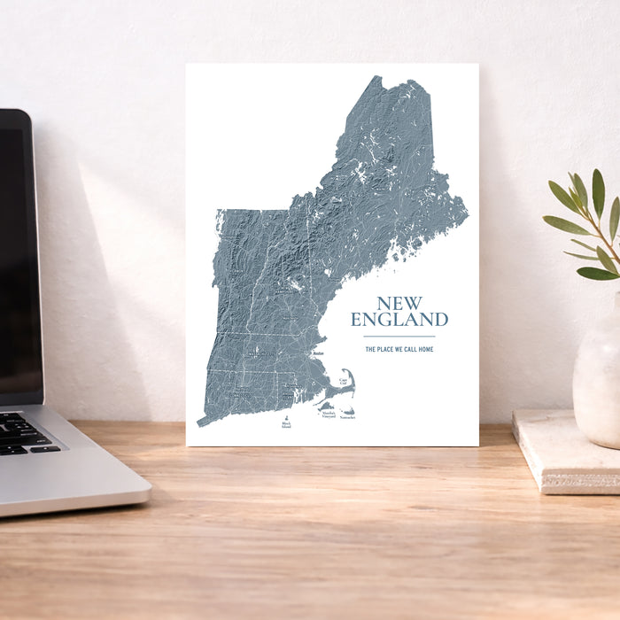 A vertical New England map art print in muted blue-gray on a white background, displayed on a wooden desk next to a laptop and a small plant in a vase.