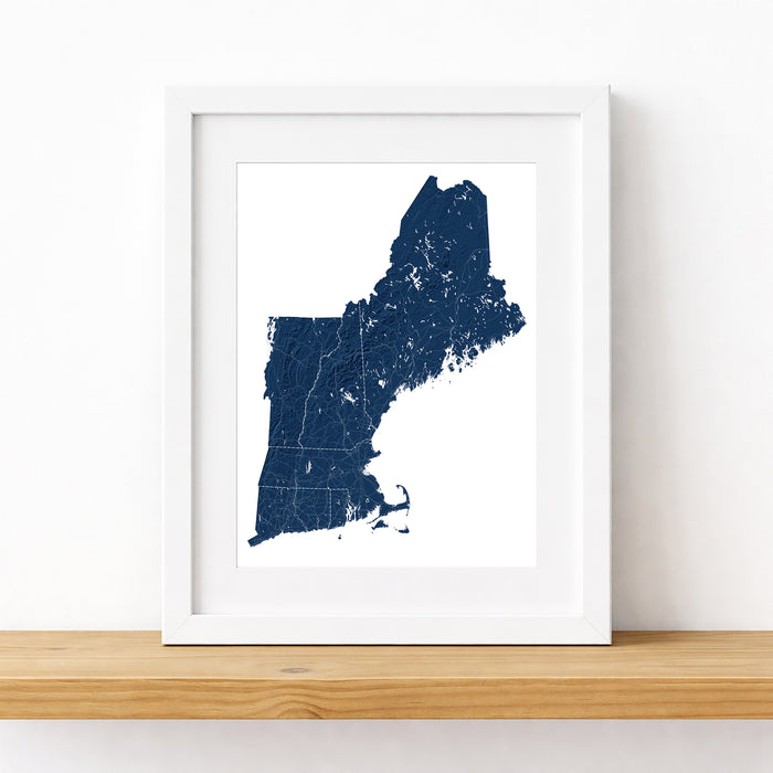 A framed state map of Maine in dark navy blue with white lines, presented in a white frame on a light wood shelf against a white wall.