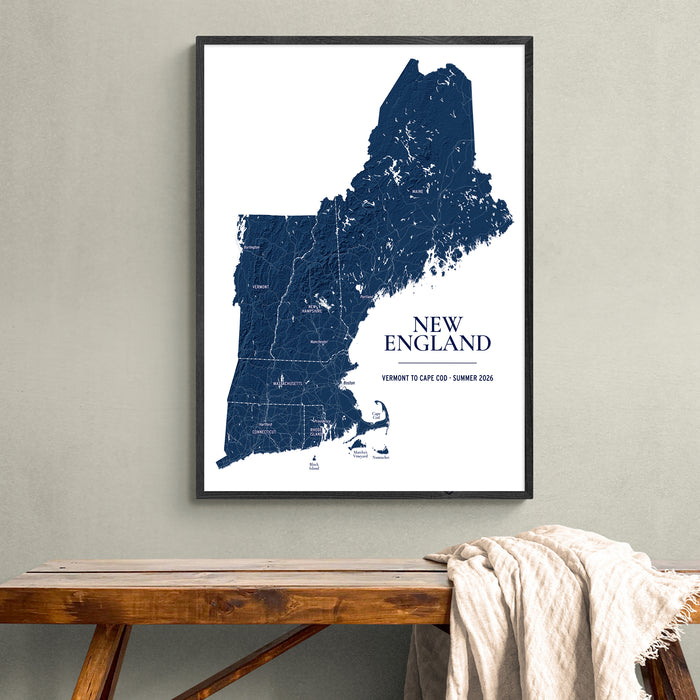 A framed New England state map print hangs on a light gray wall above a wooden bench with a draped cream throw blanket. The poster shows a dark blue map of New England with the text 'NEW ENGLAND' and smaller location text at the bottom.