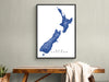 Framed map of New Zealand on a wall above a wooden table.