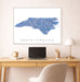 Framed map of North Carolina on a wall above a desk with a laptop and lamp.
