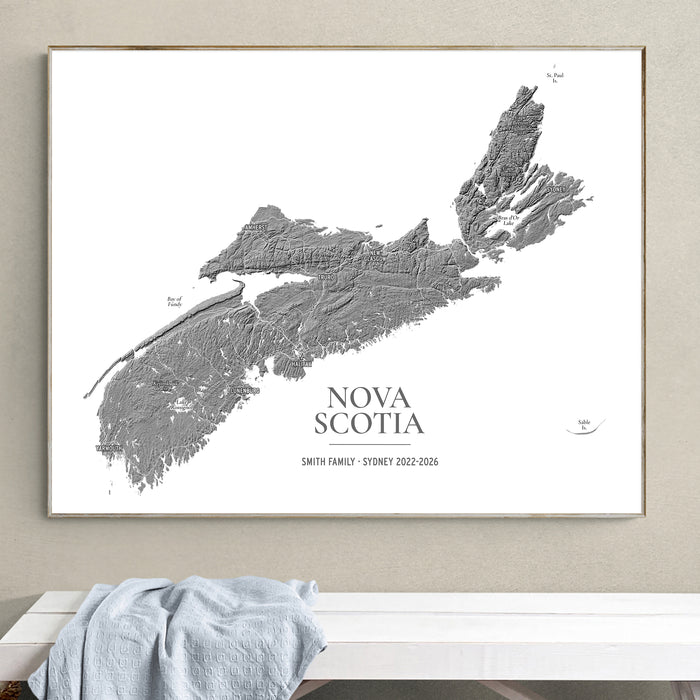 A framed custom topographic map print of Nova Scotia, featuring gray landforms on a white background with the text 'NOVA SCOTIA' and 'SMITH FAMILY - SYDNEY 2022-2026'. The framed print is displayed on a beige wall above a white bench with a light blue draped cloth.