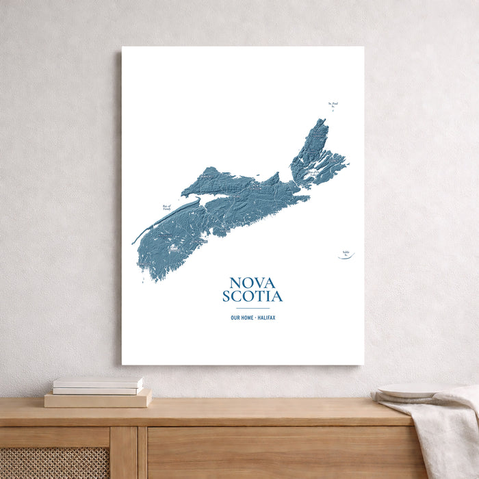A vertical white art print featuring a blue topographic map of Nova Scotia with the text 'NOVA SCOTIA' and 'OUR HOME - HALIFAX' near the bottom, displayed on a light gray wall above a wooden console table.