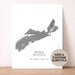 A custom Nova Scotia topographic map art print displayed on a light wood surface against a textured beige wall. The print features a detailed gray map of Nova Scotia on a white background with the text 'NOVA SCOTIA' and a custom date.
