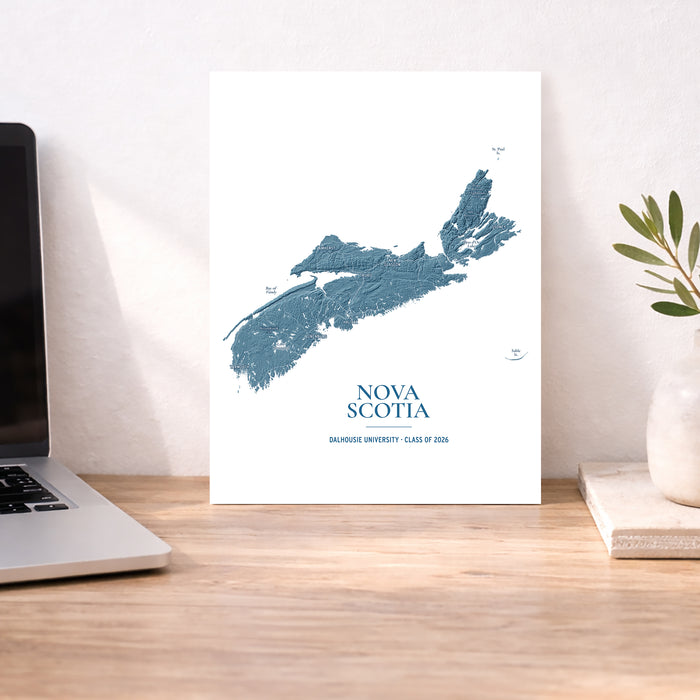 A vertical white art print featuring a blue map of Nova Scotia with the text 'NOVA SCOTIA' and 'BAIRDGE UNIVERSITY - CLASS OF 2026' at the bottom, displayed on a wooden desk next to a laptop and a small plant in a vase.