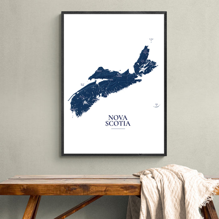 A framed Nova Scotia map art print in dark blue and white, hanging on a light gray wall above a rustic wooden bench with a draped cream throw blanket.