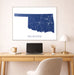 Framed map of Oklahoma on a wall above a desk with a laptop and lamp.