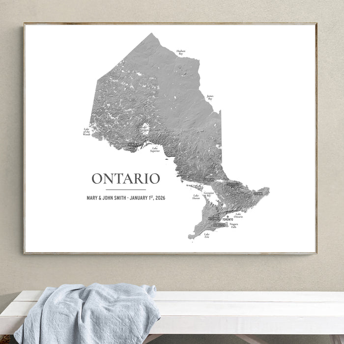 A framed monochrome Ontario map print hangs on a beige wall above a white bench with a light blue draped cloth. The print shows the outline of Ontario with labeled regions and the text 'ONTARIO' plus personalized names and date.