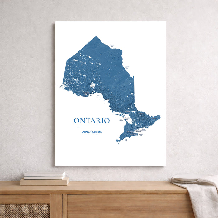 A vertical wall art print showing a blue map of Ontario, Canada, displayed on a light gray wall above a wooden console table with books and a throw blanket.