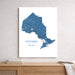 A vertical wall art print showing a blue map of Ontario, Canada, displayed on a light gray wall above a wooden console table with books and a throw blanket.