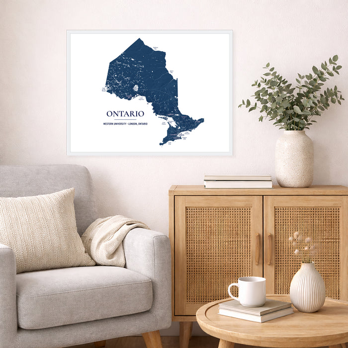 A framed wall art print showing a navy blue map of Ontario, Canada, styled in a modern living room setting next to a gray armchair and a wooden cabinet.