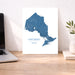 A vertical white art print featuring a blue map of Ontario, Canada, styled on a wooden desk next to a laptop and a small potted plant.
