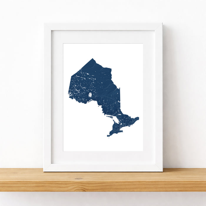 A framed art print featuring a dark navy blue map silhouette of New York State with a distressed texture, displayed on a light wood shelf against a white wall.