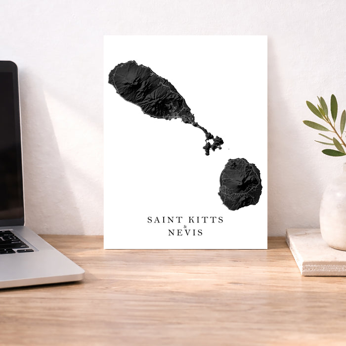 Map of Saint Kitts and Nevis on a white background displayed on a desk with a laptop and plant.