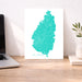 Teal map of St. Lucia on a white background, displayed on a wooden desk with a laptop and plant.
