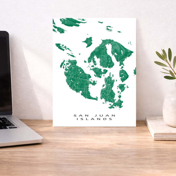 Map of San Juan Islands on a white background, displayed on a wooden desk with a laptop and plant.