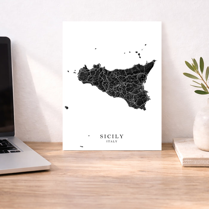 Map of Sicily on a white background next to a laptop on a wooden desk.