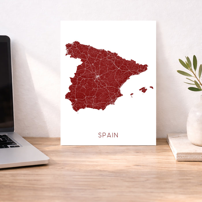 Map of Spain on a white background next to a laptop on a wooden desk.