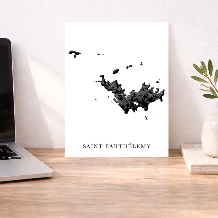Map of Saint Barthelemy on a white background next to a laptop on a wooden desk.