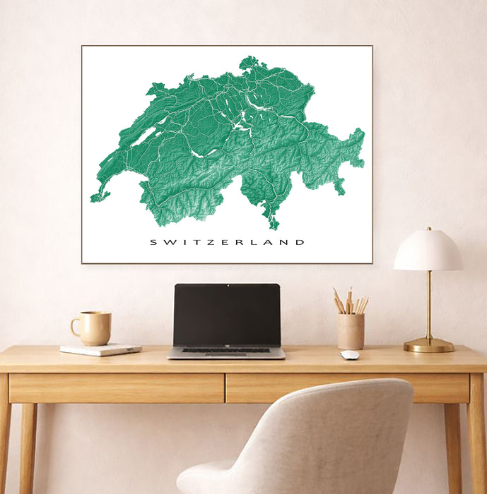 Framed map of Switzerland on a wall above a desk with a laptop and lamp.