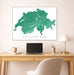 Framed map of Switzerland on a wall above a desk with a laptop and lamp.