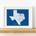 Framed map of Texas on a blue background with white roads, set against a white wall.
