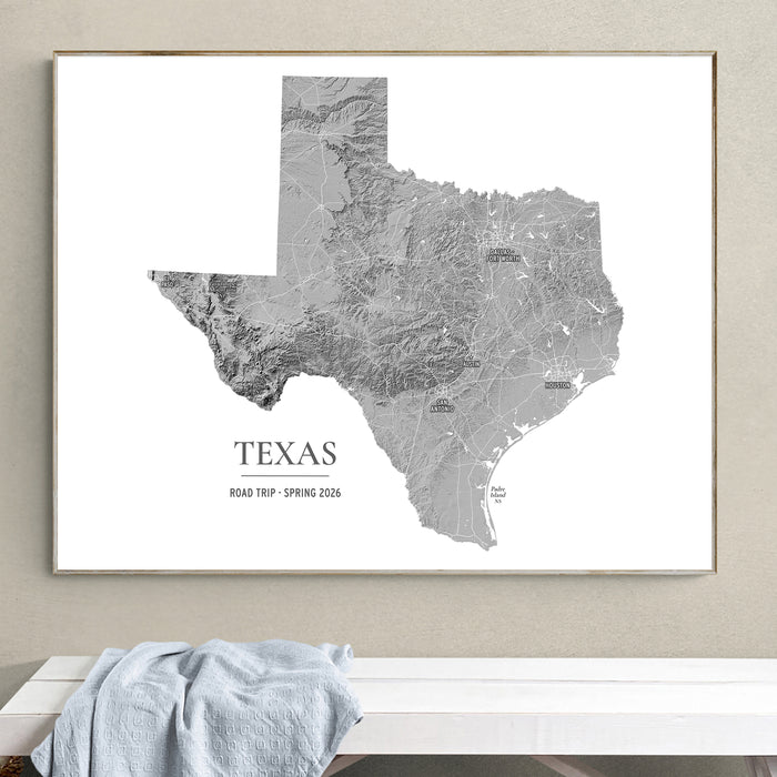 A framed gray topographic map print of Texas with the text 'TEXAS ROAD TRIP - SPRING 2026' below the map, displayed on a beige wall above a white bench with a light blue draped cloth.