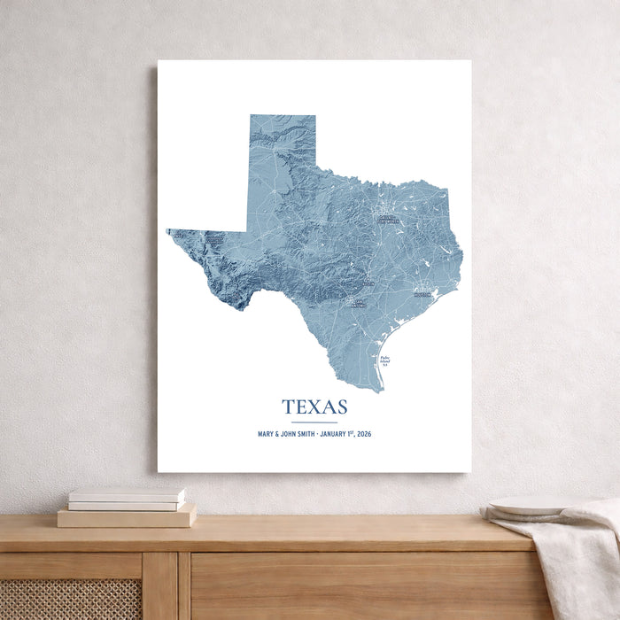 A vertical canvas print of a blue topographic map of Texas, displayed on a light gray textured wall above a wooden console table with books and a throw blanket.