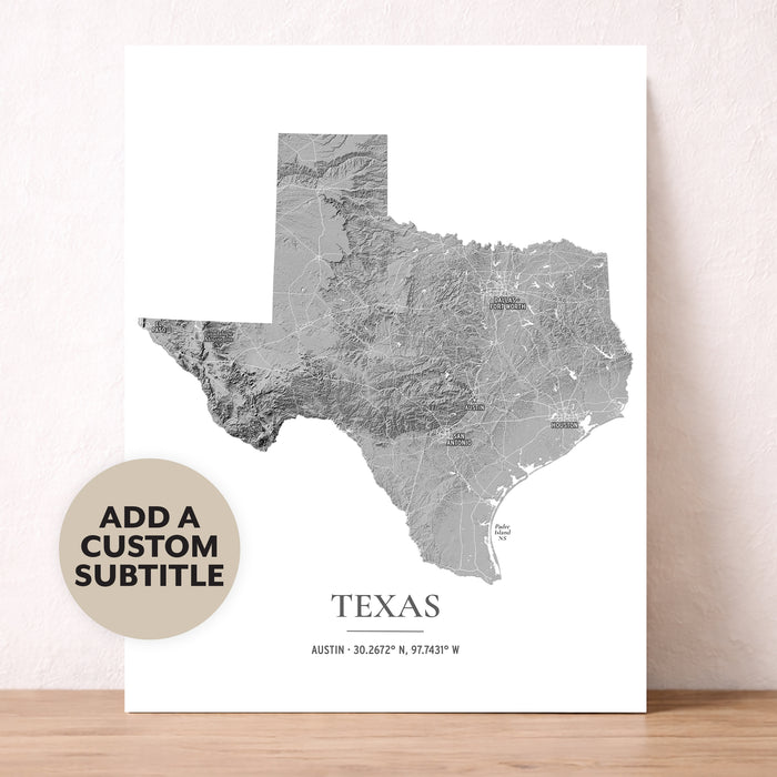 A vertical white art print featuring a gray shaded-topography map of Texas with the state name and coordinates printed below. The print is leaning against a light beige wall on a wooden surface, and a circular badge in the lower left says 'ADD A CUSTOM SUBTITLE'.