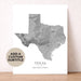 A vertical white art print featuring a gray shaded-topography map of Texas with the state name and coordinates printed below. The print is leaning against a light beige wall on a wooden surface, and a circular badge in the lower left says 'ADD A CUSTOM SUBTITLE'.