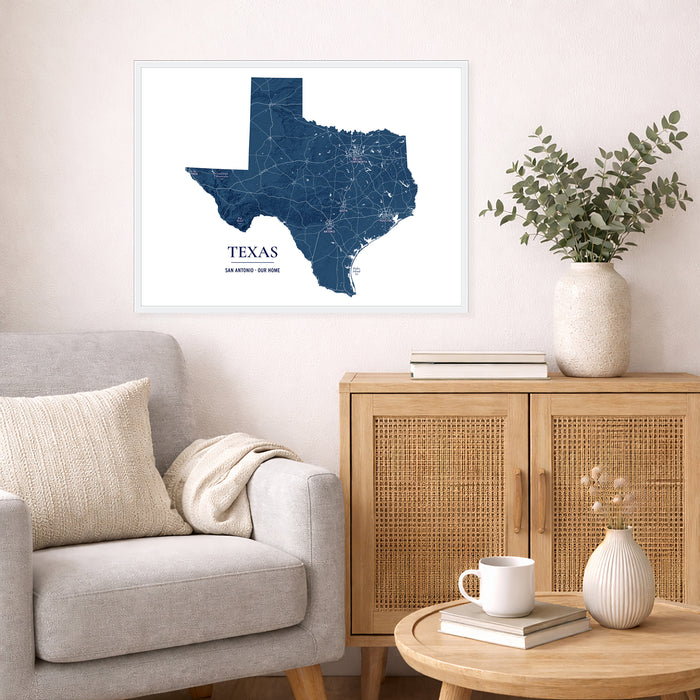 A framed Texas state map print in navy blue and white hangs on a light beige wall above a gray armchair and a wooden cabinet with a vase of greenery in a styled living room.