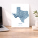 A vertical map print of Texas in shades of blue and gray on a white background, displayed on a wooden desk next to a laptop and a small plant in a vase.