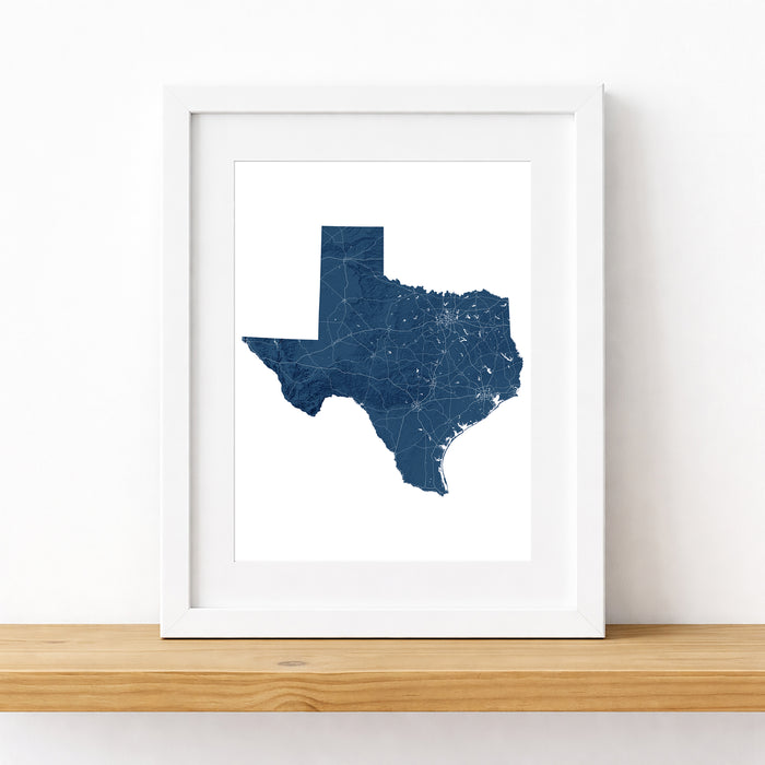 A framed art print featuring a dark blue map of Texas with white city highlights, displayed in a white frame on a light wood shelf against a white wall.